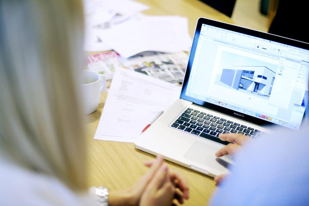 Et bilde av et bord med en mac med digital plantegning av hus, med dokumenter og kaffekopp rundt. 