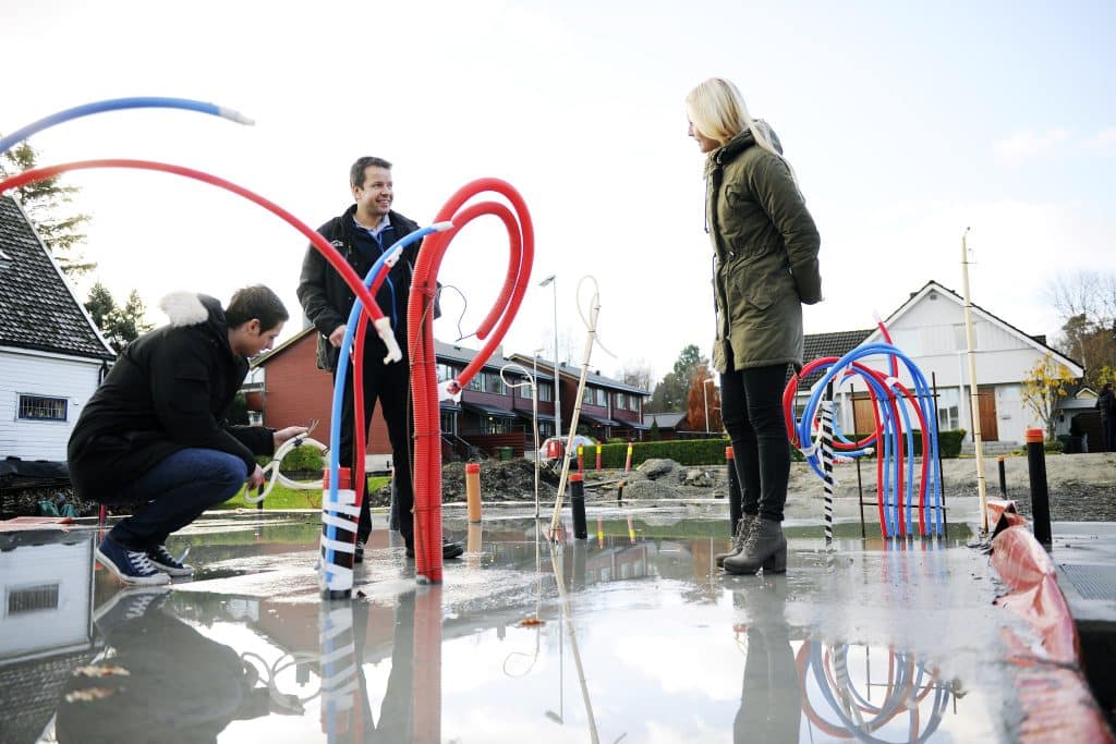 Ungt par og byggeleder står på ferdig grunnmur med plastrør og ledninger stikkende opp.