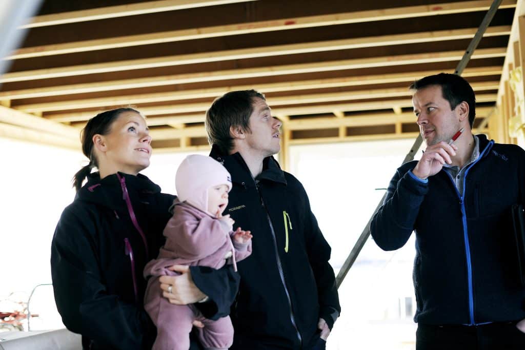 Byggeleder prater med mann mens kvinne holder baby og ser opp smilende. 