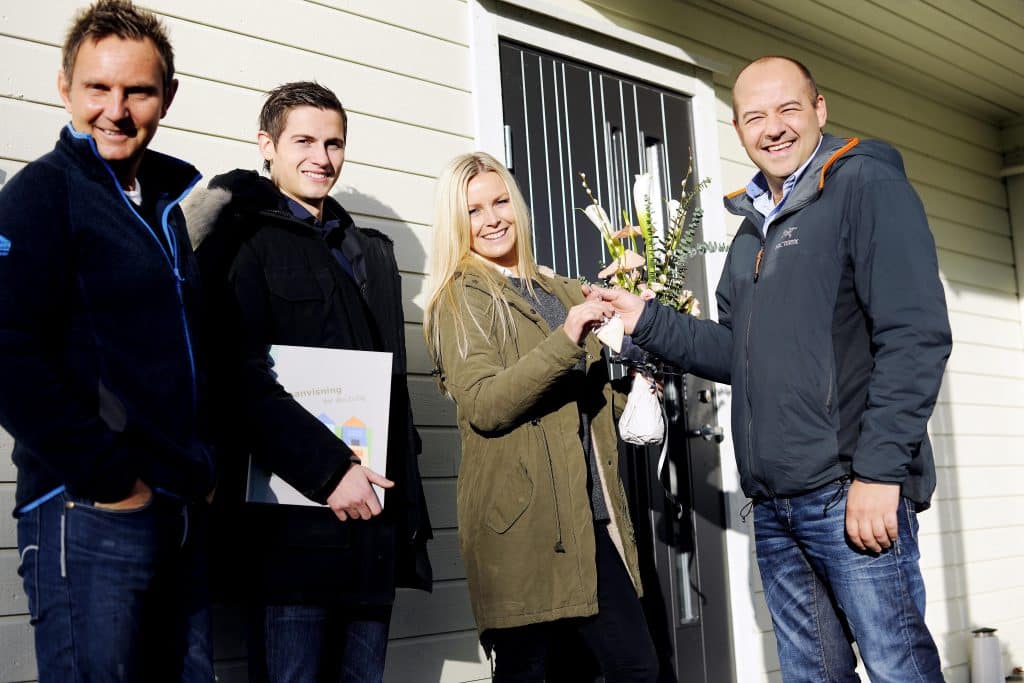 Ungt par mottar husnøkkel og gratulasjon av boligbyggeren og byggelederen utenfor dør til nytt hus. 