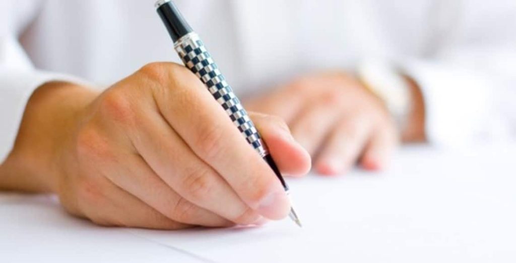 To hender som en signerer med en kulepenn og den andre hånden hviler på bordet. 
